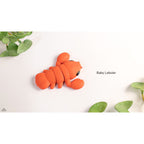 Baby Lobster articulated fidget toy — official Zou3D render, angled overhead with plant lifestyle props, orange colourway